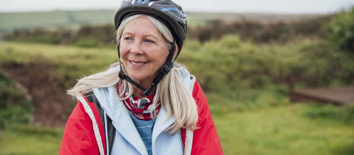 A shot of a senior woman standing wearing a cycling helmet and holding her bike looking at the scenery smiling.
